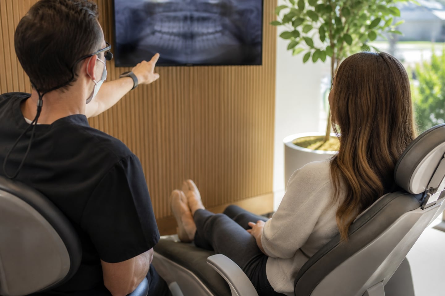 A dentist pointing at a screen while explaining a treatment plan to a seated patient.